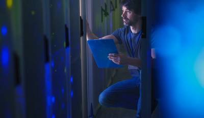 Blue-tinged view of a man in jeans and T-shirt referring to a clipboard while checking an IT data centre.